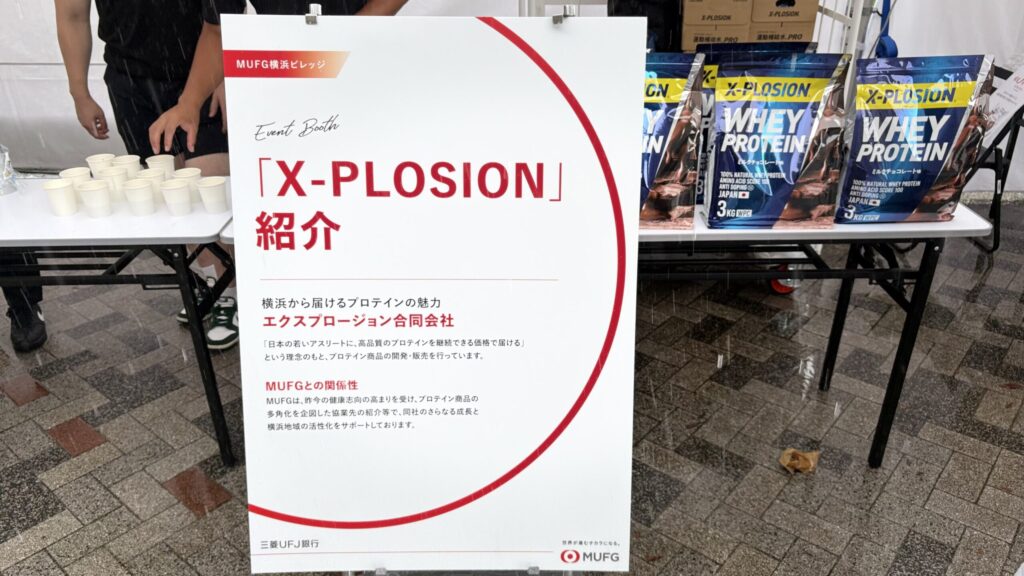 X-PLOSIONの紹介パネルと、試飲用の紙コップが並ぶテーブル