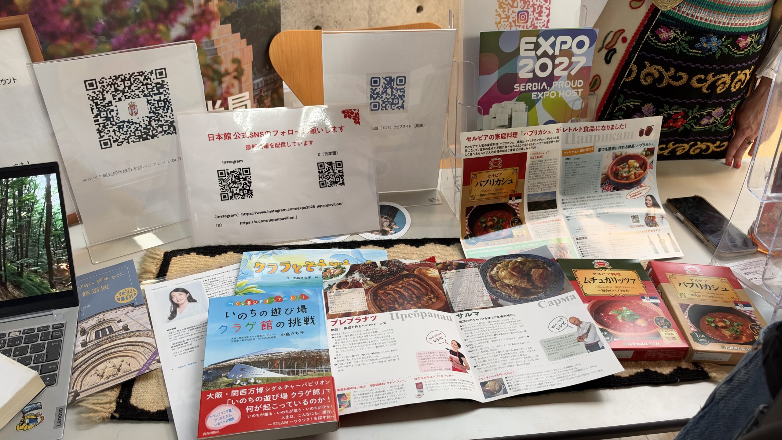 日本館のSNS案内、クラゲ館の本、セルビアのパンフレットとレトルト食品が並ぶブース