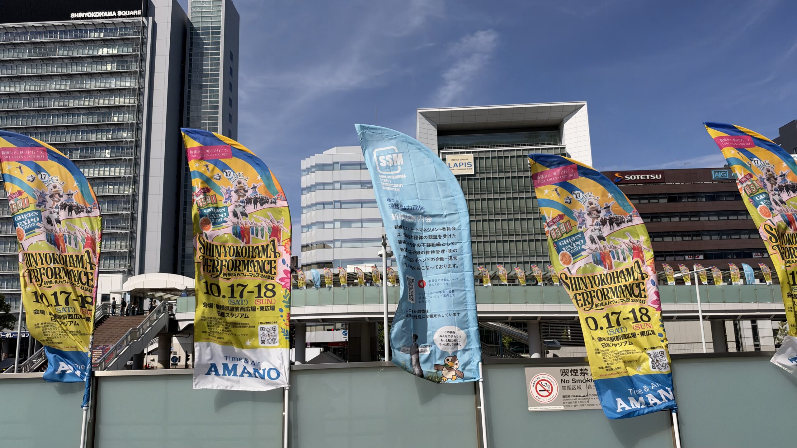 新横浜駅前の歩道橋に並ぶ新横浜パフォーマンス2026の旗と、新横浜ストリートマネジメント委員会の旗