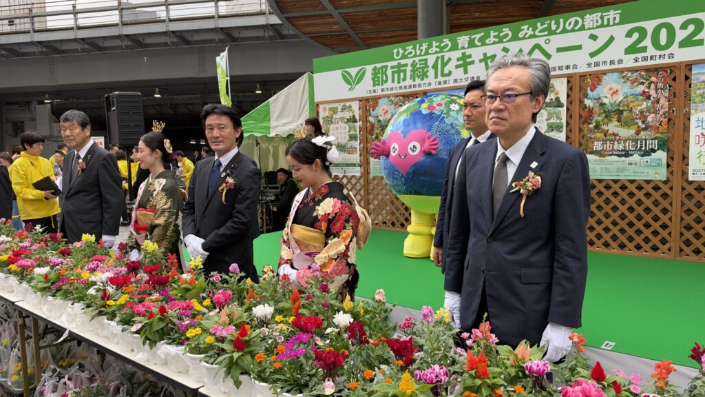 花鉢が並ぶテーブルの前に立つ出席者と、ステージ上のトゥンクトゥンク